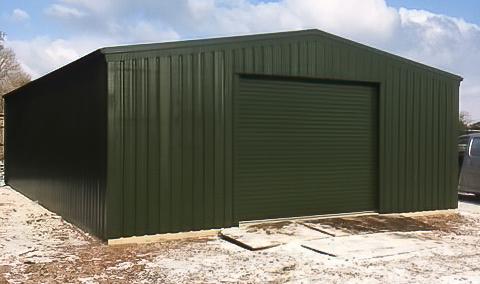 A wide-angle shot of a large green steel warehouse with a central roller shutter door sitting on a concrete foundation in a snowy field.