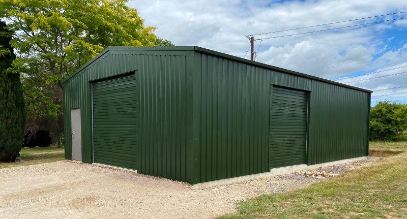 Corner view of a dark green steel building showing a front roller door, a side roller door, and a personnel door on a gravel foundation.